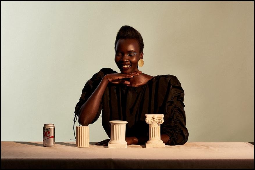 Smiling Black model poses with small column sculptures and a Diet Coke can on a table in front of an off-white background. 