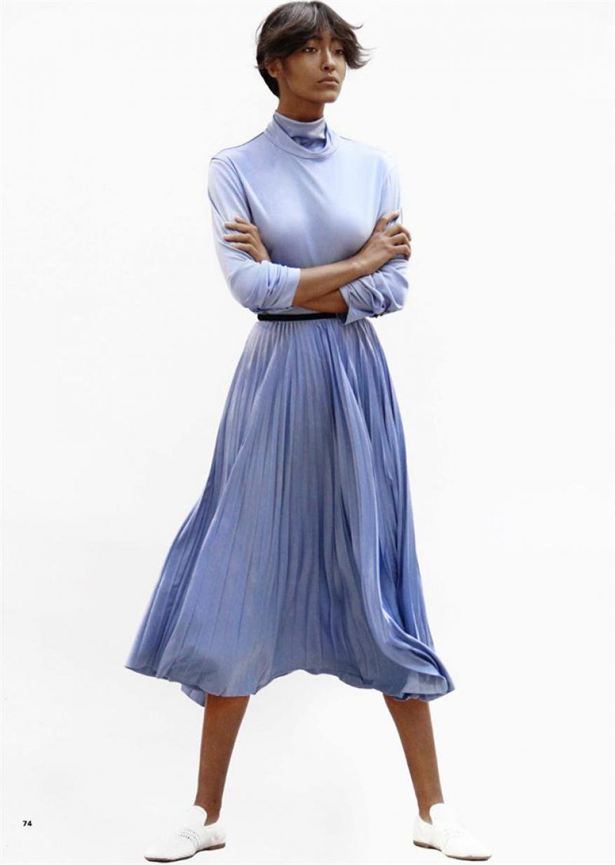 A woman in a light blue dress with long sleeves and a pleated skirt is standing in a white photo studio with arms crossed.