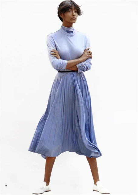 A woman in a light blue dress with long sleeves and a pleated skirt is standing in a white photo studio with arms crossed.