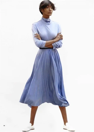 A woman in a light blue dress with long sleeves and a pleated skirt is standing in a white photo studio with arms crossed.
