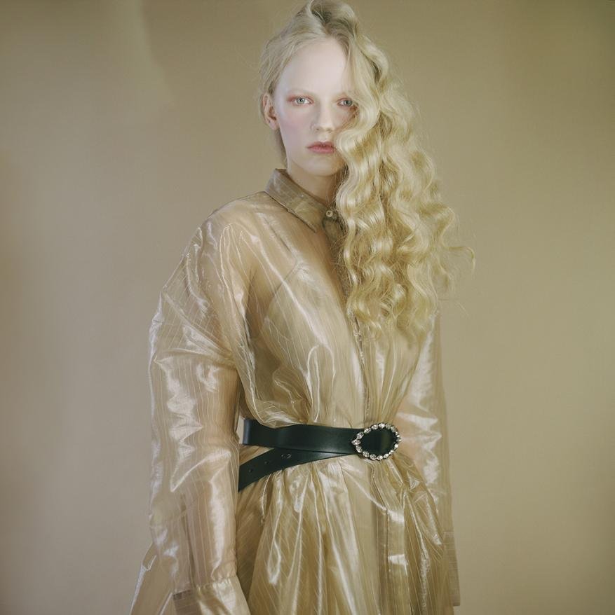 Fashion portrait of a blonde woman wearing a transparent overcoat cinched with a black belt against a neutral backdrop in a studio setting.