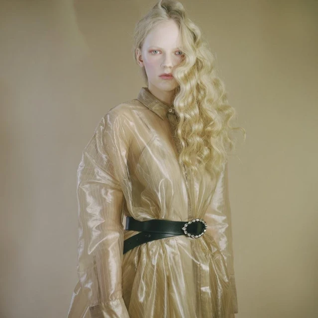Fashion portrait of a blonde woman wearing a transparent overcoat cinched with a black belt against a neutral backdrop in a studio setting.