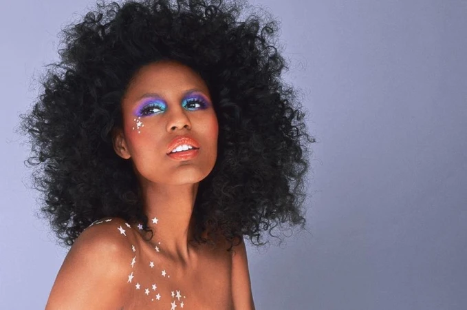 Black woman with voluminous curly dark hair, vibrant eye makeup, and star stickers on her face and shoulder against a grey background