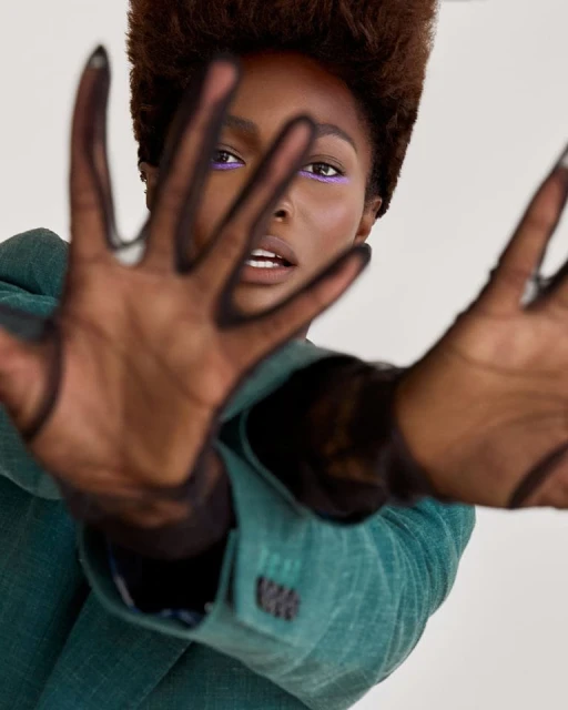 Portrait of a woman with an Afro hairstyle, wearing a green suit and sheer gloves, posing with her hands up in front of her face.