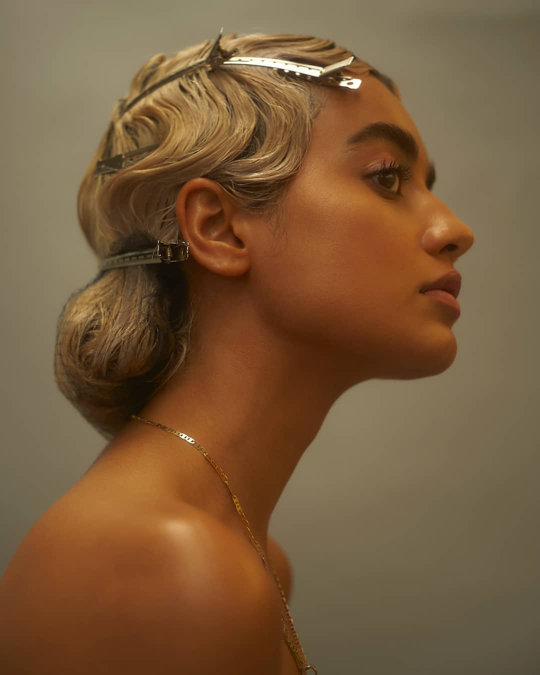 Profile shot of an adult female with light skin and blonde wavy hair styled with metal clips, wearing a gold chain necklace indoors.