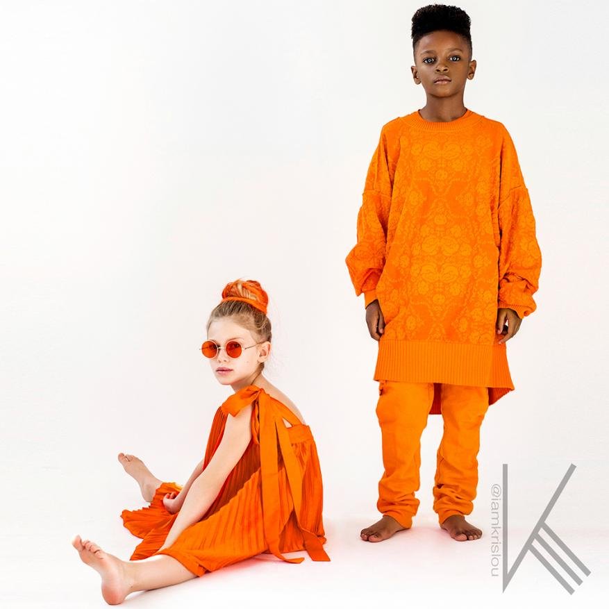 Two children, a boy and a girl, are posing in orange outfits against a white background in a fashion style setting. The boy is standing, and the girl is seated.