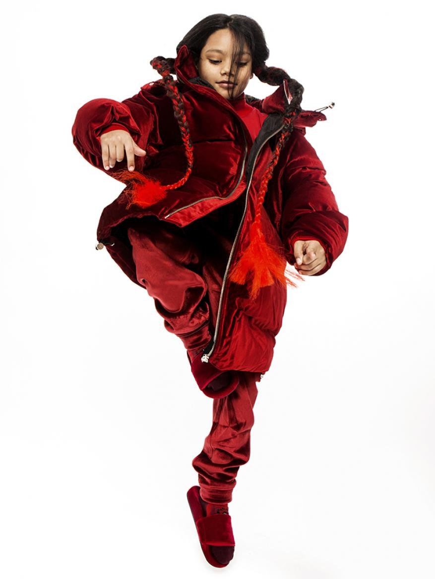 Young girl strikes an energetic pose in a red velvet puffer jacket and matching pants against a bright white background, braids and movement.