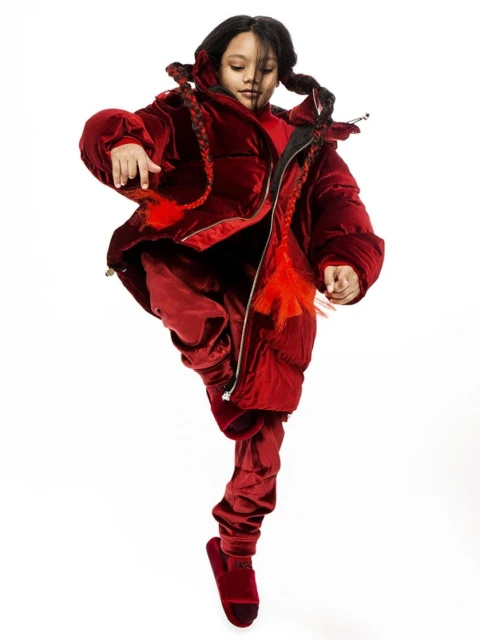 Young girl strikes an energetic pose in a red velvet puffer jacket and matching pants against a bright white background, braids and movement.
