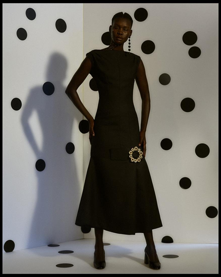 Elegant woman in a black dress stands in a studio with a white backdrop decorated with black polka dots. High fashion portrait.