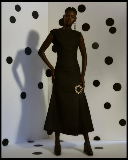 Elegant woman in a black dress stands in a studio with a white backdrop decorated with black polka dots. High fashion portrait.