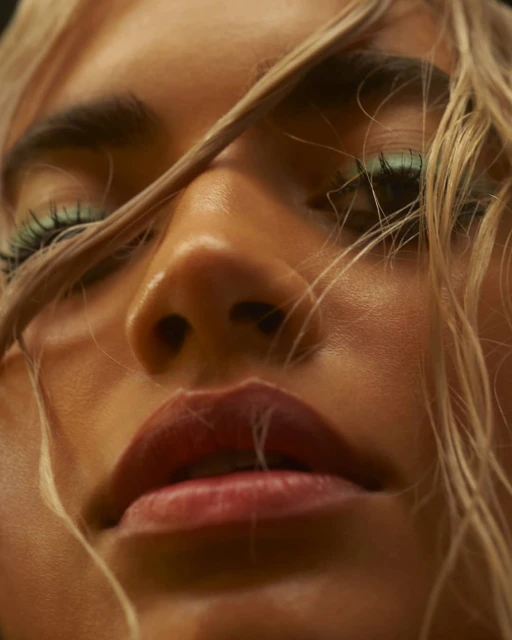 Close-up portrait of a woman with blonde hair and green eye makeup, with focus on facial details like eyes, nose, and lips.