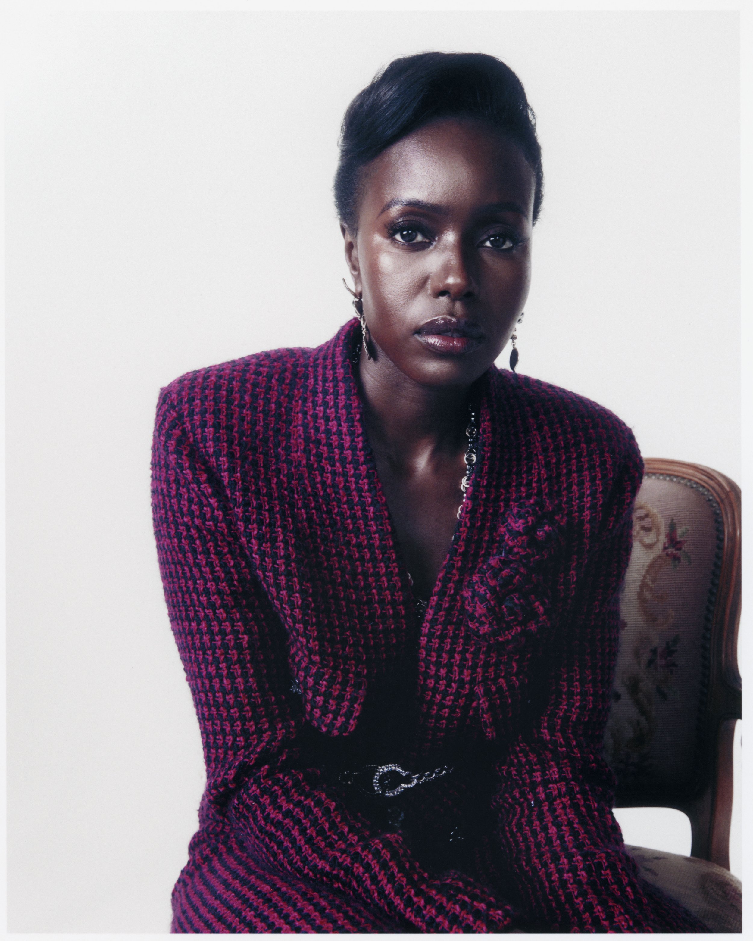 Elegant portrait of a Black woman in a textured purple blazer and a black belt, seated against a bright white backdrop, fashion editorial.
