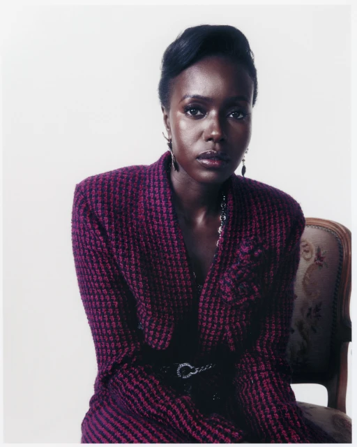 Elegant portrait of a Black woman in a textured purple blazer and a black belt, seated against a bright white backdrop, fashion editorial.