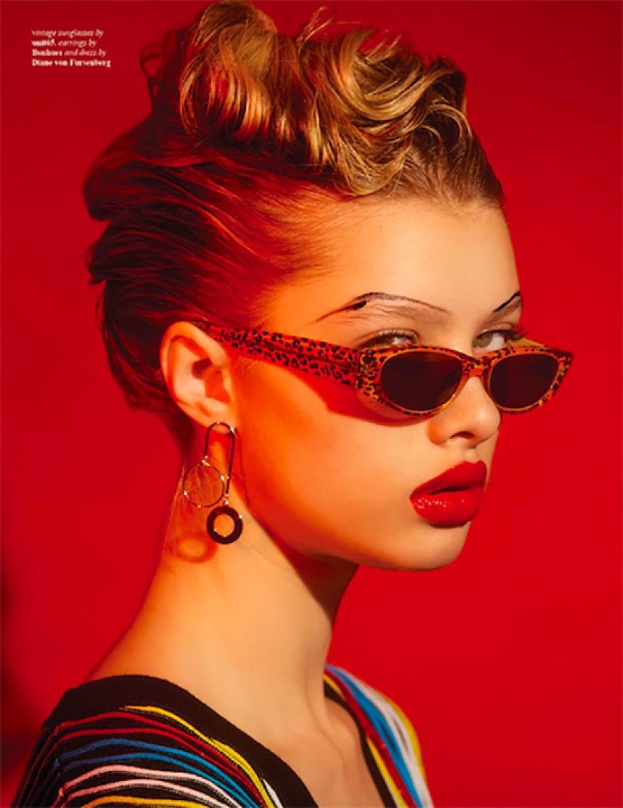 Portrait of a fair-skinned woman with blonde updo, red lipstick, and eyewear against a solid red backdrop in a beauty-focused shot.