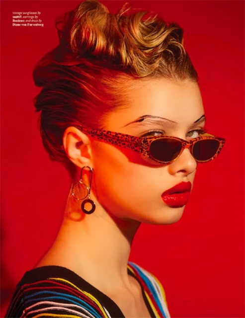 Portrait of a fair-skinned woman with blonde updo, red lipstick, and eyewear against a solid red backdrop in a beauty-focused shot.