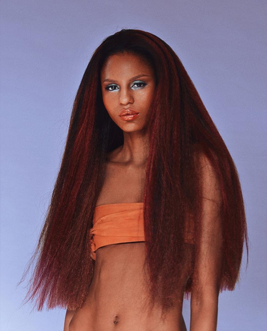 A young woman with long, voluminous red-brown hair, wearing an orange top against a light purple background, showcases a beauty and fashion shot.