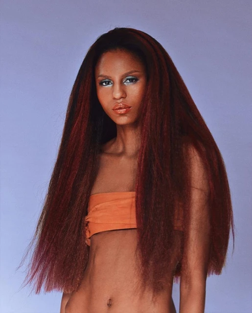 A young woman with long, voluminous red-brown hair, wearing an orange top against a light purple background, showcases a beauty and fashion shot.