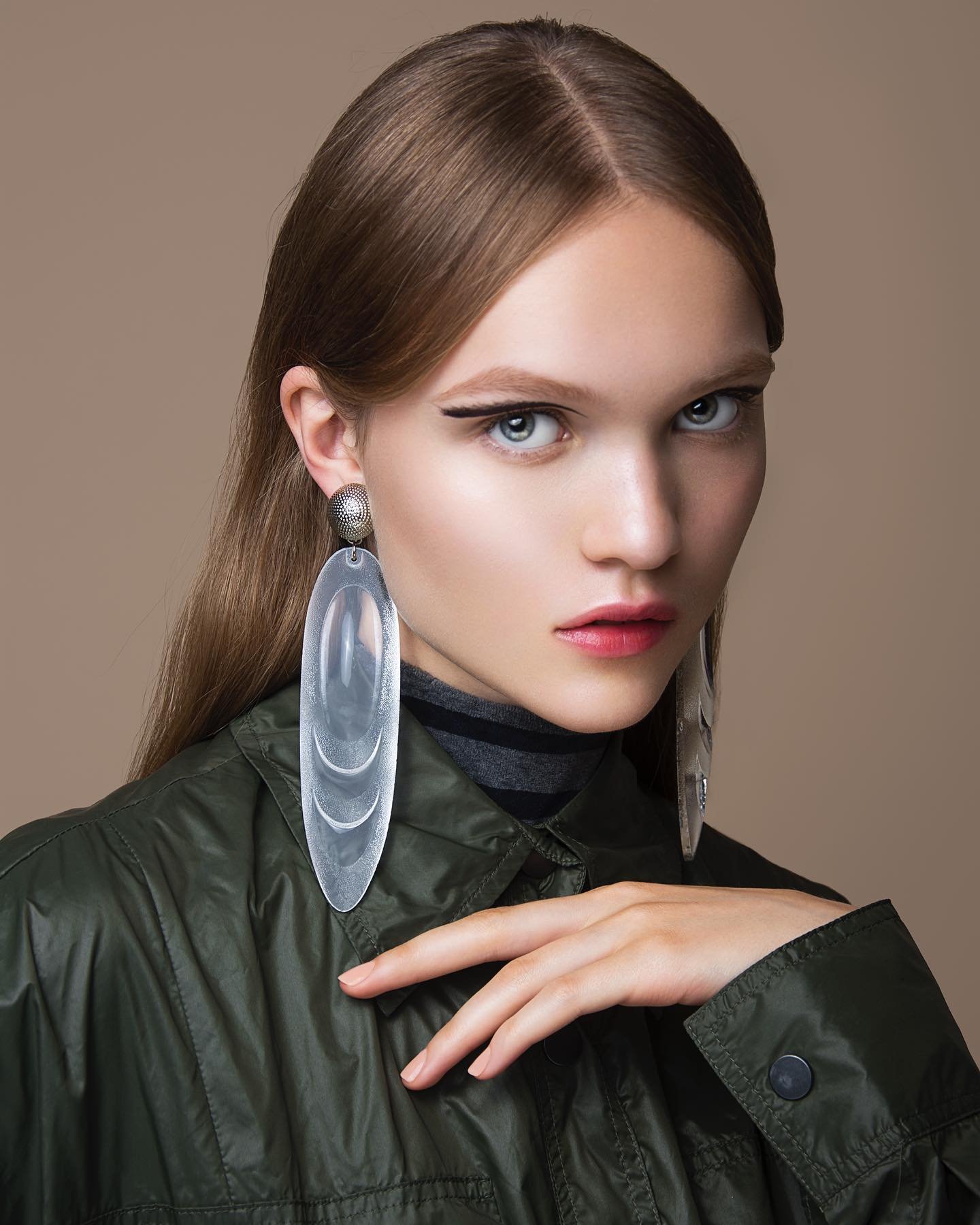 Close-up of a young adult female with long straight hair, wearing a green jacket, large earrings, and makeup against a beige background.