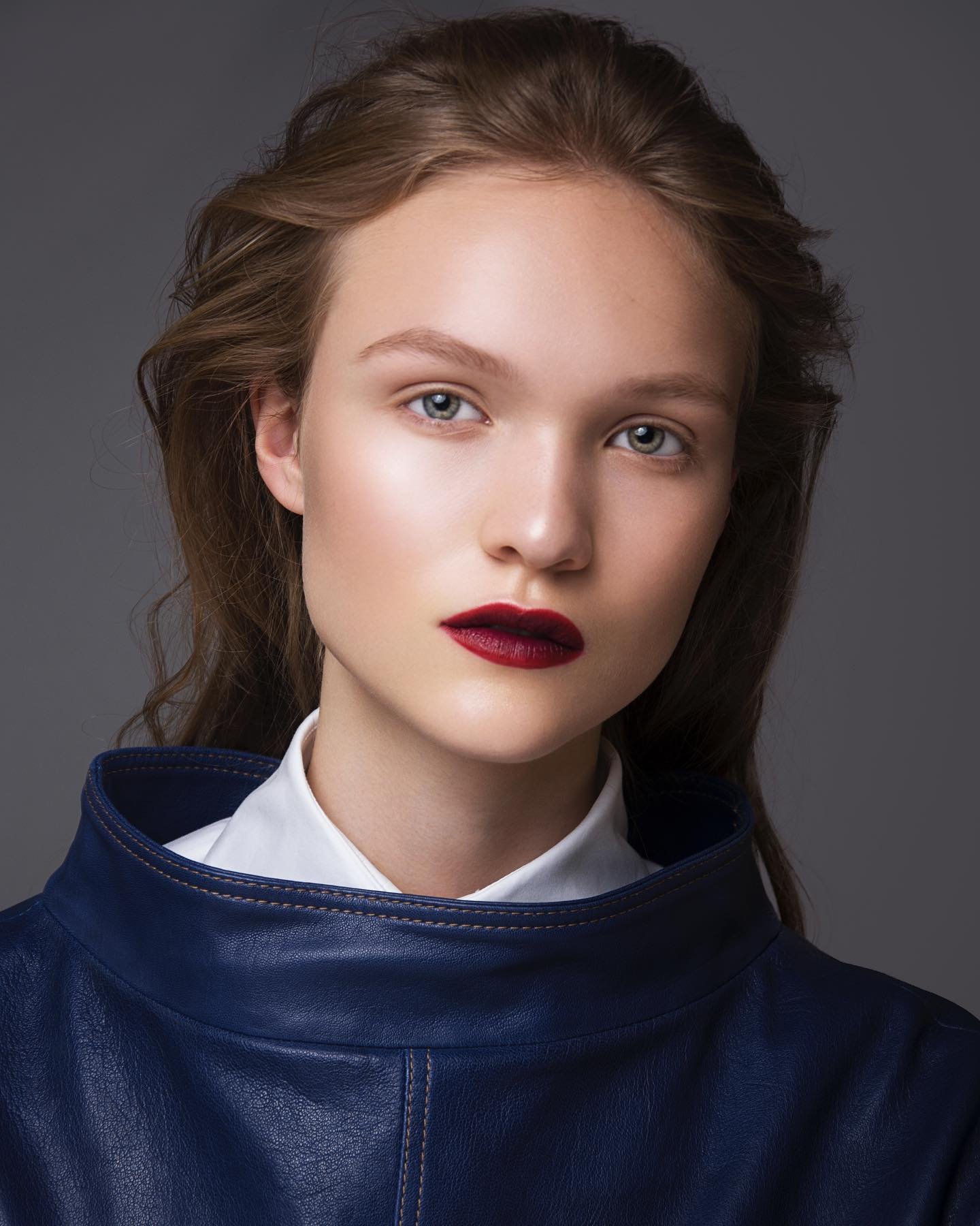 Close-up portrait of a woman with dark red lipstick and long brown hair wearing a blue high-neck top against a gray background.