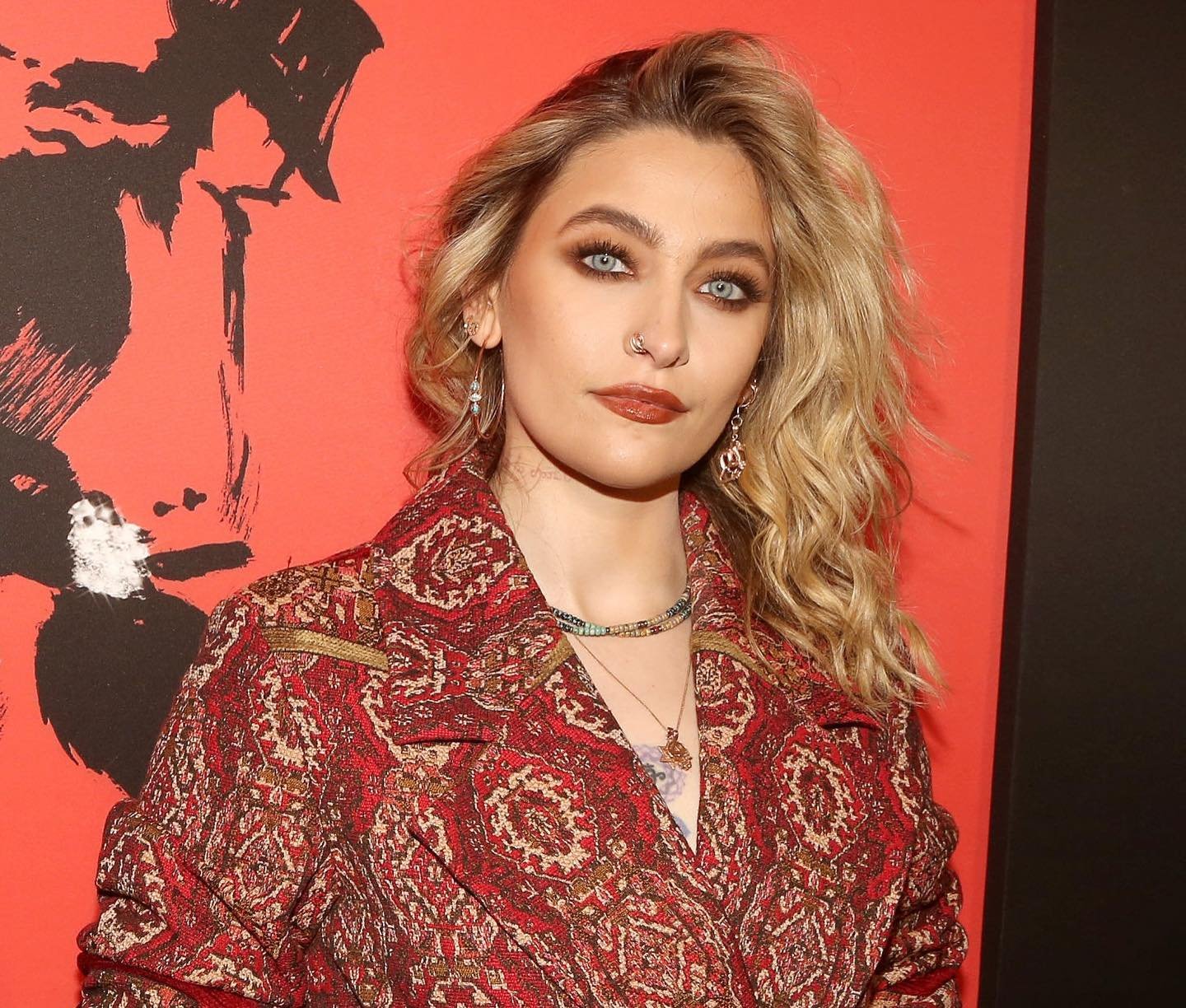 Portrait of Paris Jackson with wavy blonde hair, wearing a patterned red jacket, indoors against an abstract red and black background.
