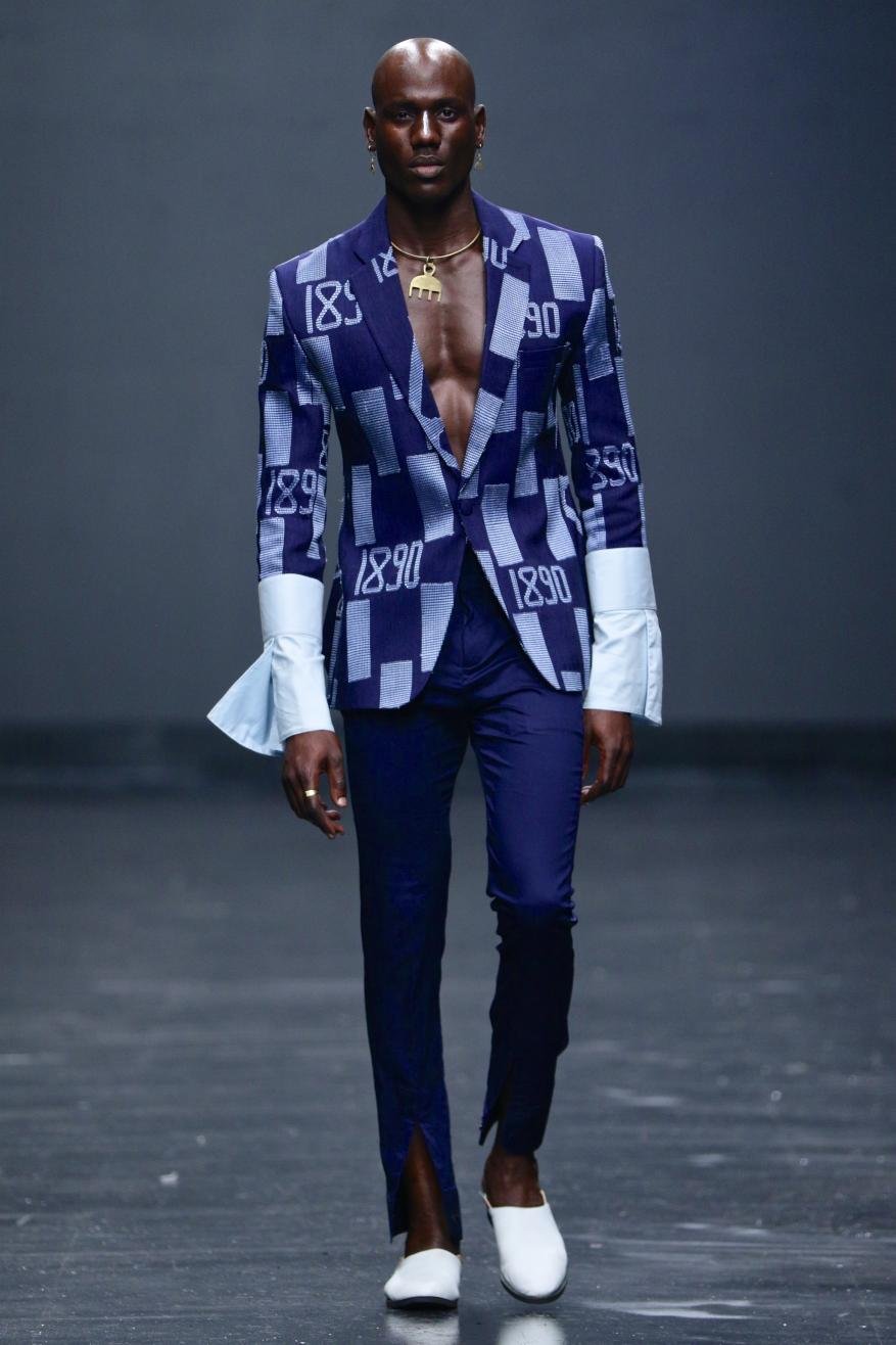 Fashion model on runway wearing an open blue patterned blazer with white cuffs, blue trousers with front slits, and white shoes.