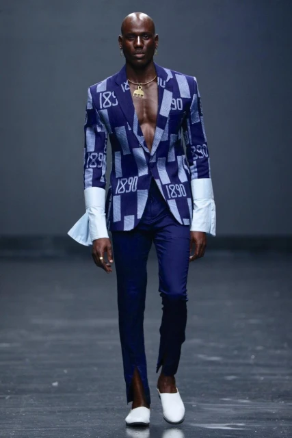 Fashion model on runway wearing an open blue patterned blazer with white cuffs, blue trousers with front slits, and white shoes.
