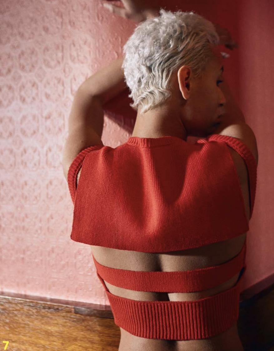From behind, a person with short white hair poses against a pink wall, wearing a red backless crop top with strappy details.