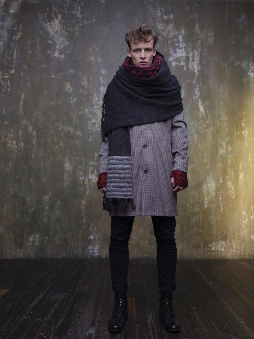 A fair-skinned man stands wearing a long coat, jeans, boots, and a large dark scarf in front of a textured wall in a studio setting.