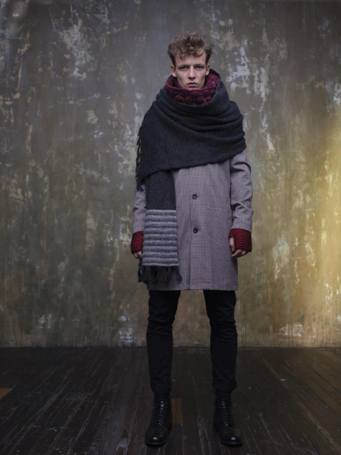 A fair-skinned man stands wearing a long coat, jeans, boots, and a large dark scarf in front of a textured wall in a studio setting.
