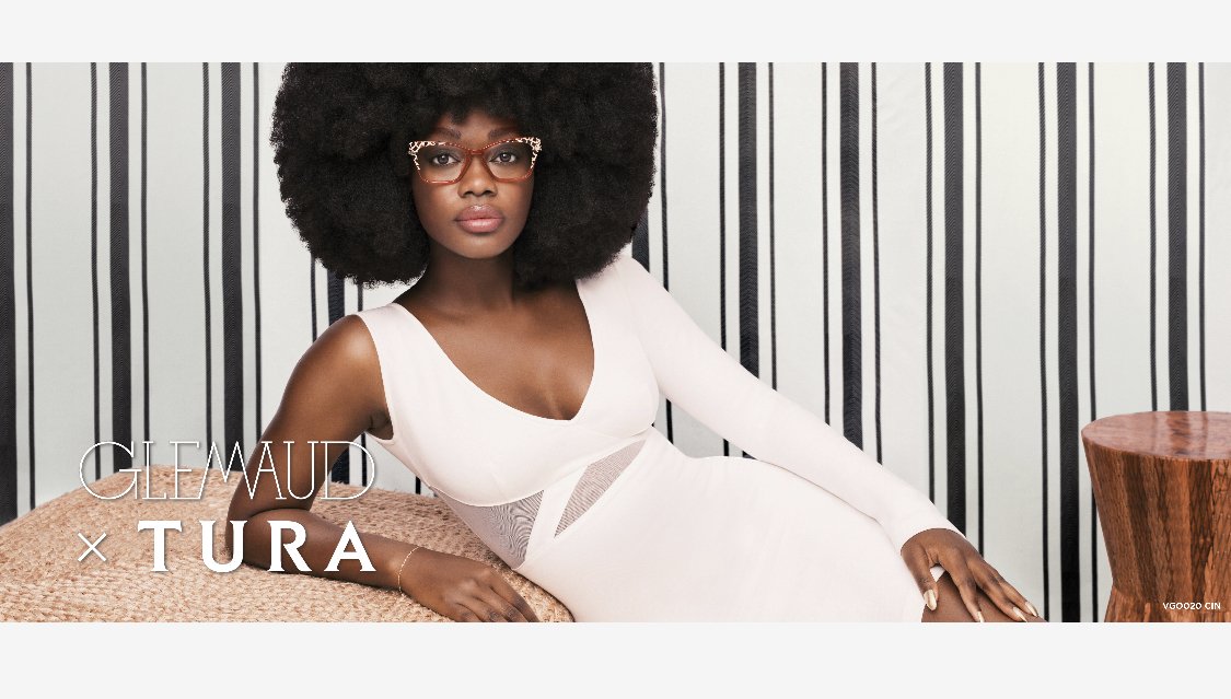 Fashion shot of a Black woman wearing eyeglasses and a light pink dress, posing in front of a striped backdrop and wood stool.
