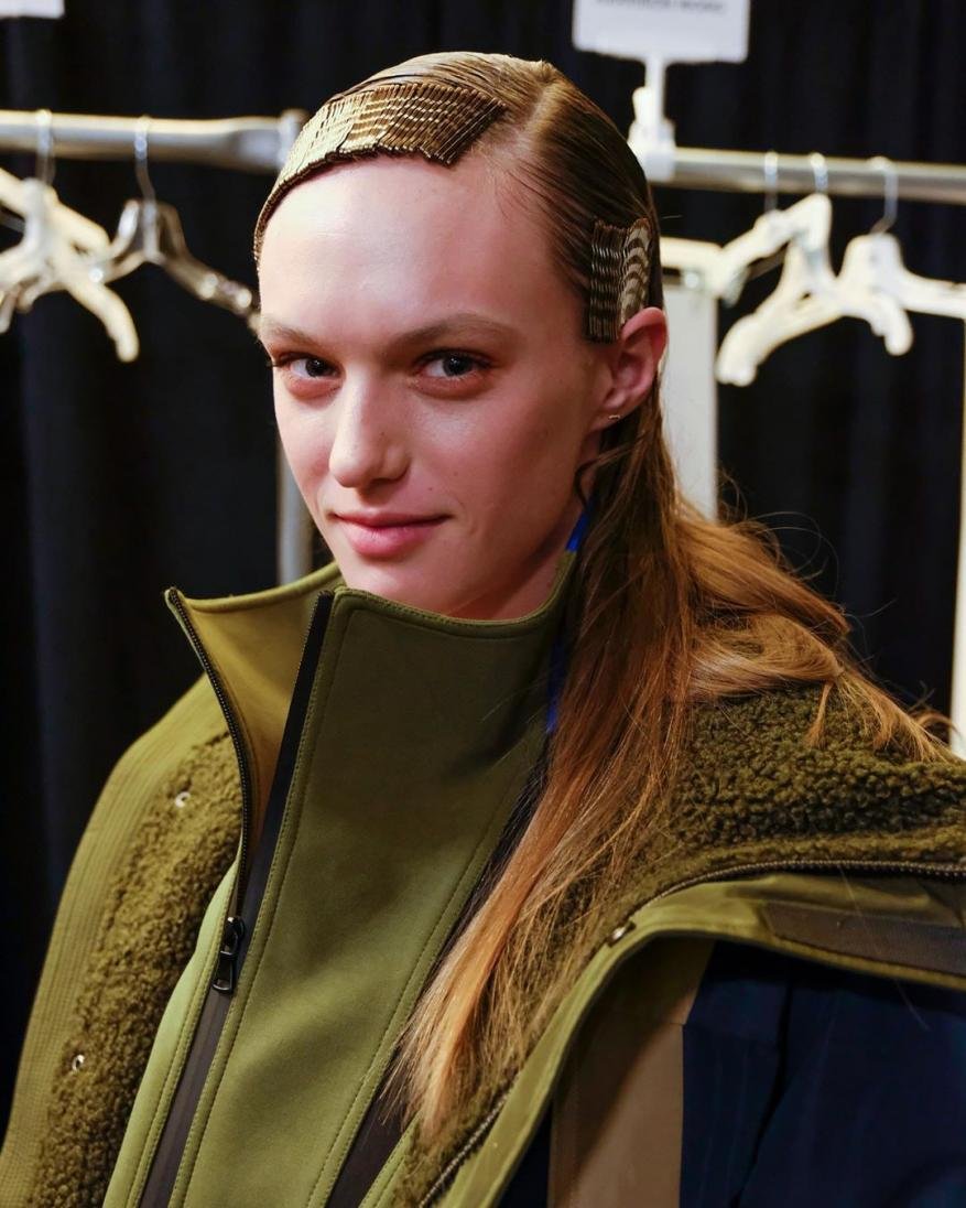 A young woman poses indoors wearing an olive green jacket, with her long brown hair styled with gold decorative hair clips.