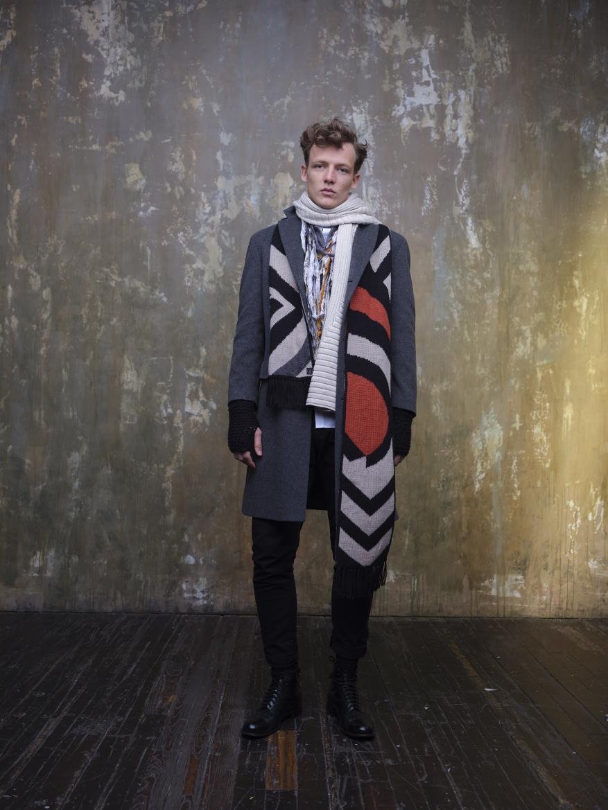 Fashionable young man in a gray coat and patterned scarf stands indoors against a mottled, textured wall with dark wood flooring.