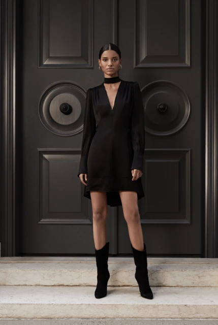 Woman in long-sleeved black dress and black boots poses in front of a dark grey ornate door on light grey marble steps.