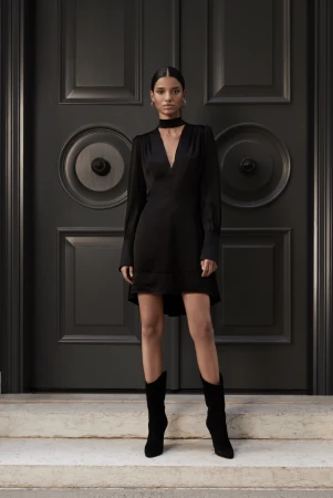 Woman in long-sleeved black dress and black boots poses in front of a dark grey ornate door on light grey marble steps.