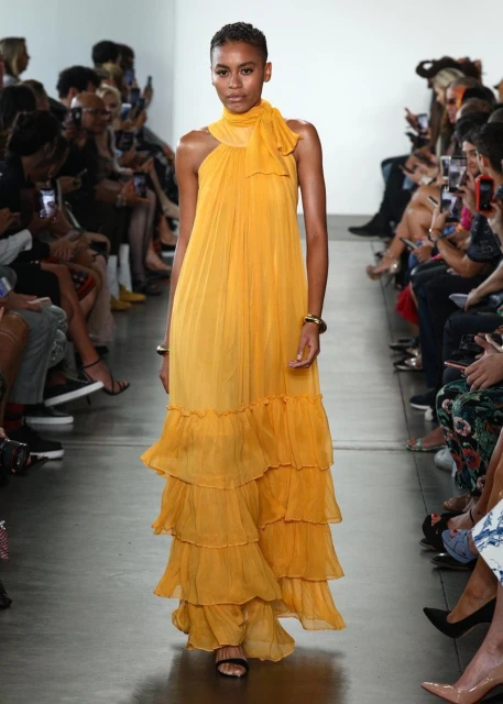 Runway fashion show featuring a model in a long, flowing yellow dress with tiered ruffles, accessorized with jewelry, walking on a white runway.