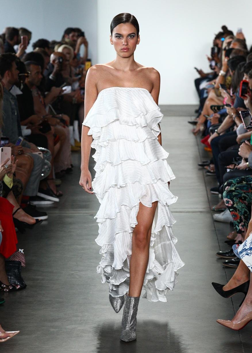Runway model wearing a white ruffle dress and silver boots, walking during a fashion show, with spectators in the background.