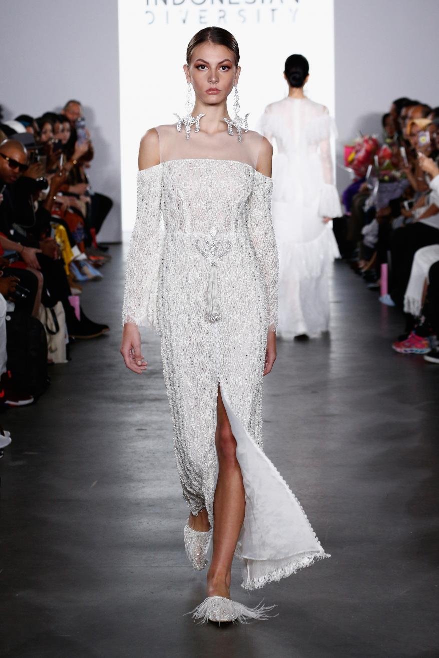 Runway show featuring a model wearing a long, white, beaded dress with off-the-shoulder sleeves at Indonesia Diversity fashion event.