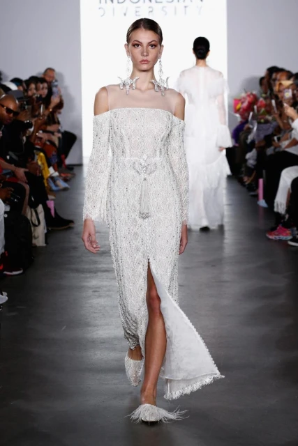 Runway show featuring a model wearing a long, white, beaded dress with off-the-shoulder sleeves at Indonesia Diversity fashion event.