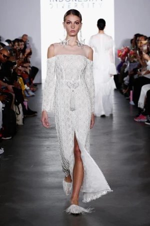 Runway show featuring a model wearing a long, white, beaded dress with off-the-shoulder sleeves at Indonesia Diversity fashion event.