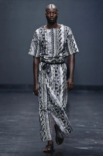 Bald Black male model walks runway in a patterned long shirt and sandals, medium shot, dark background, studio lighting, fashion photography.