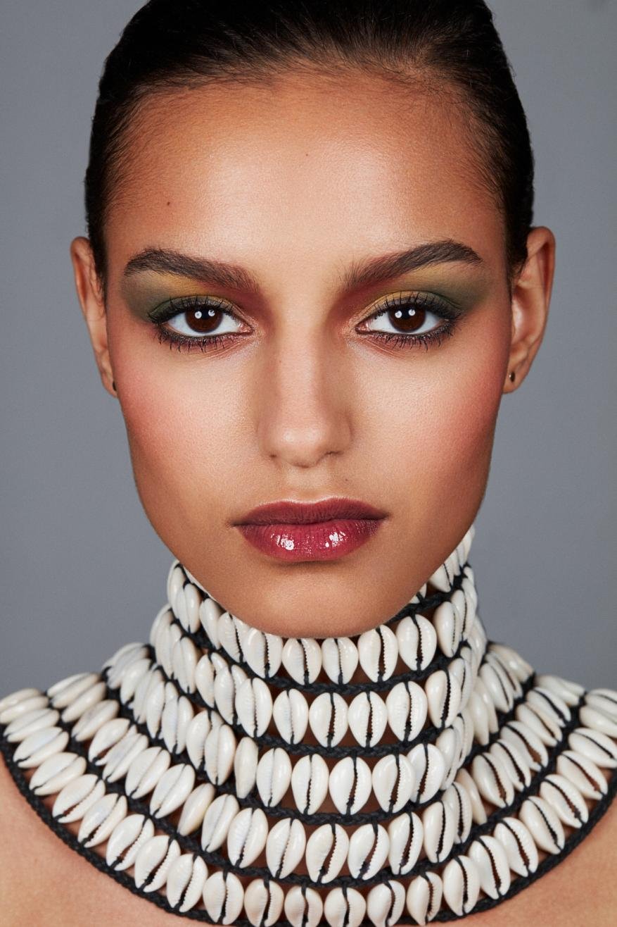 Close-up portrait of a woman with dramatic makeup and a cowrie shell necklace against a gray background in a beauty photography style.