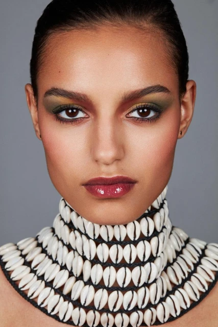 Close-up portrait of a woman with dramatic makeup and a cowrie shell necklace against a gray background in a beauty photography style.