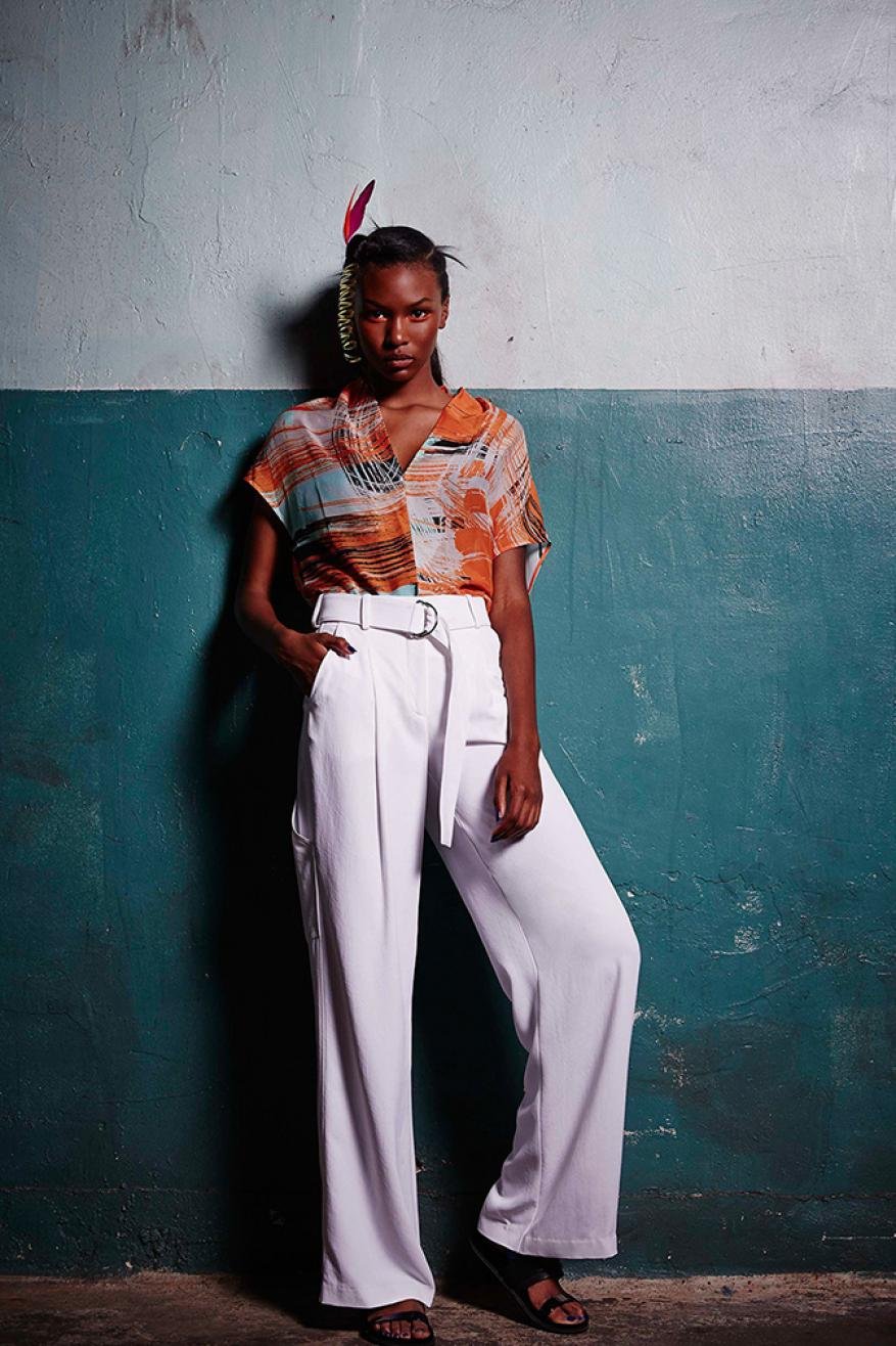 Elegant Black woman in patterned top and white belted pants poses against a teal wall, showcasing fashion and style in a studio setting.