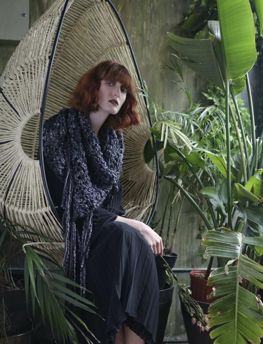 A red-haired woman sits in a hanging chair in a lush, green indoor space, wearing a dark dress and a patterned gray shawl with long fringes.