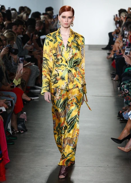 A runway model presents a yellow patterned silk suit during a fashion show, with an audience in the background holding up phones.
