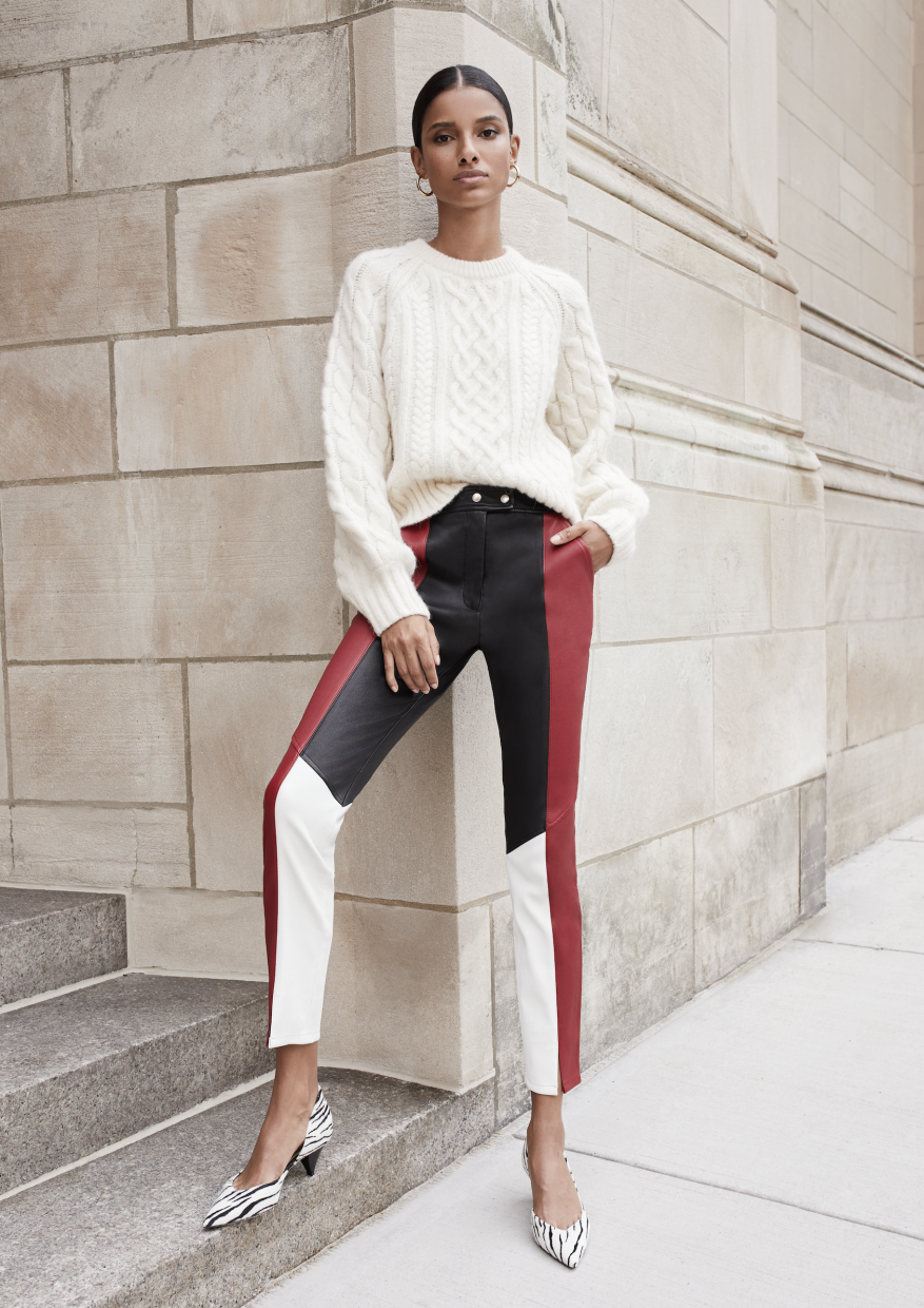 An adult woman poses in front of a textured wall wearing a cream sweater, color-blocked leather pants, and zebra-print high heels.