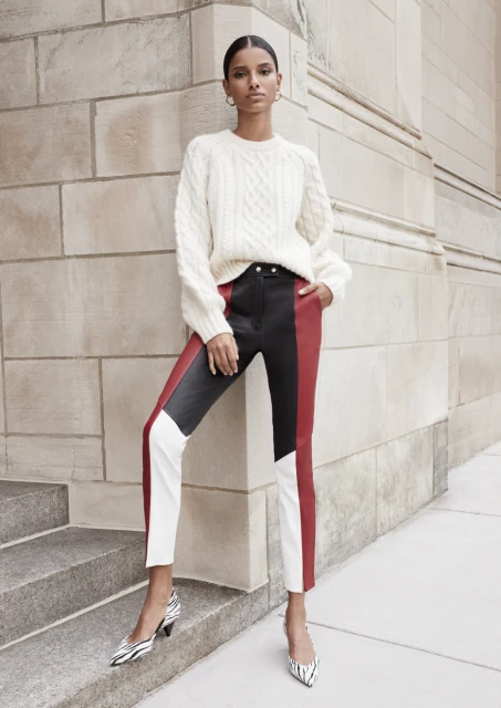 An adult woman poses in front of a textured wall wearing a cream sweater, color-blocked leather pants, and zebra-print high heels.