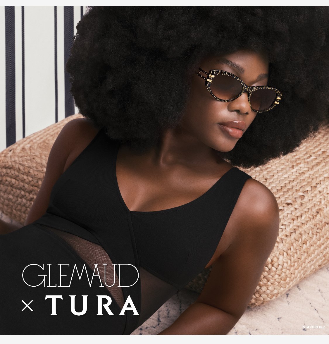 A Black woman models sunglasses while reclining in a black swimsuit. She is resting on a woven cushion in front of striped wallpaper.