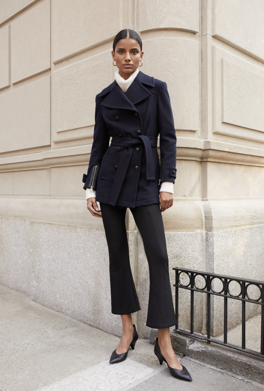 Brunette female fashion model poses in a navy belted jacket, black pants, and pointed black heels against an architectural wall.