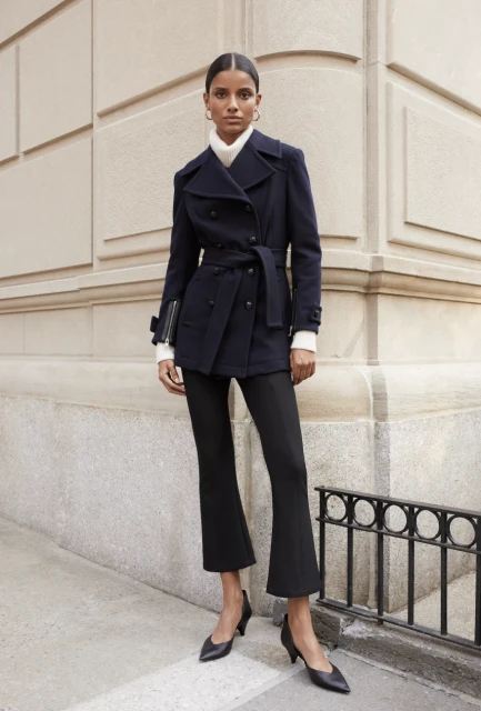 Brunette female fashion model poses in a navy belted jacket, black pants, and pointed black heels against an architectural wall.